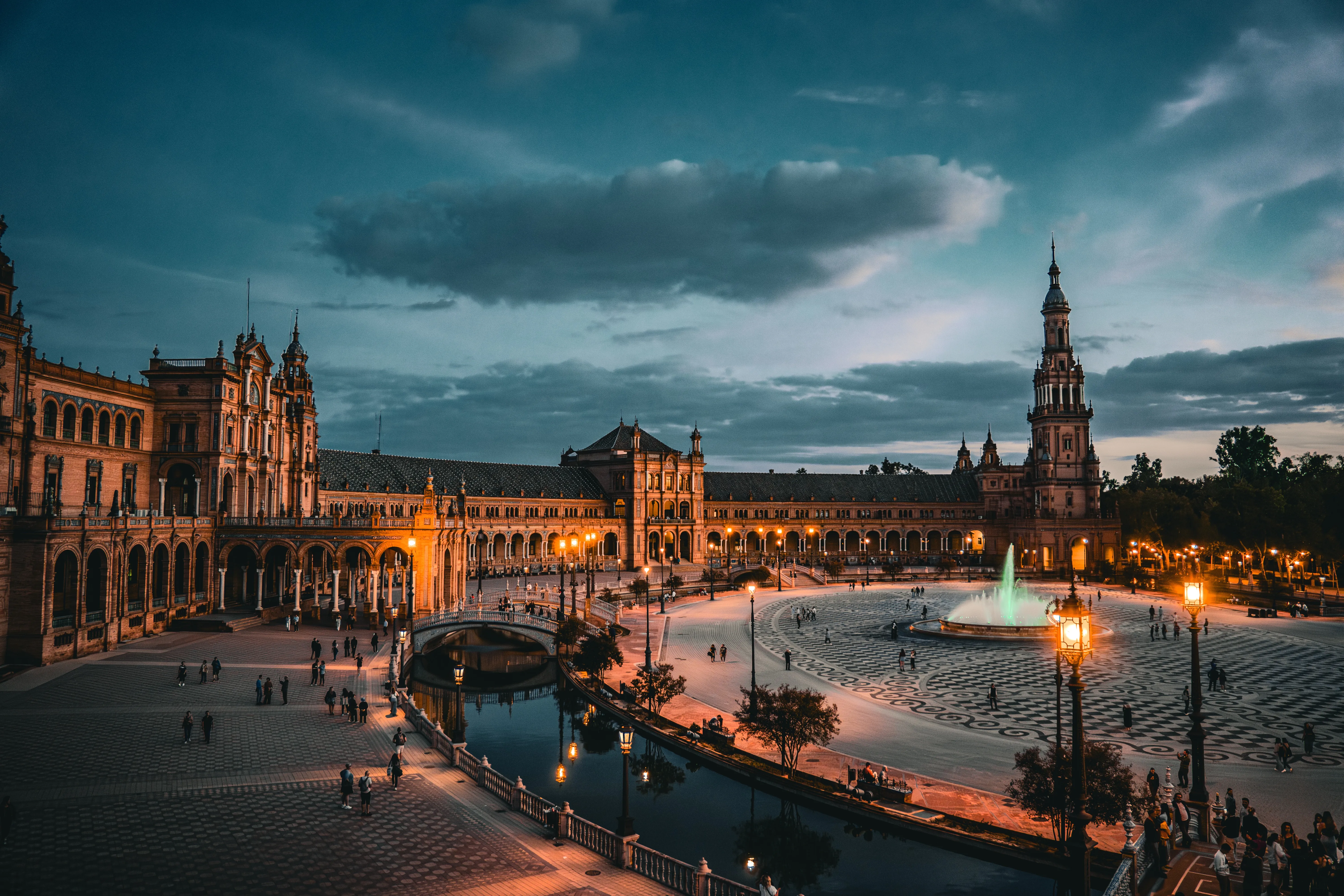 Illuminated Spanish plaza at blue hour beneath dark clouds.
