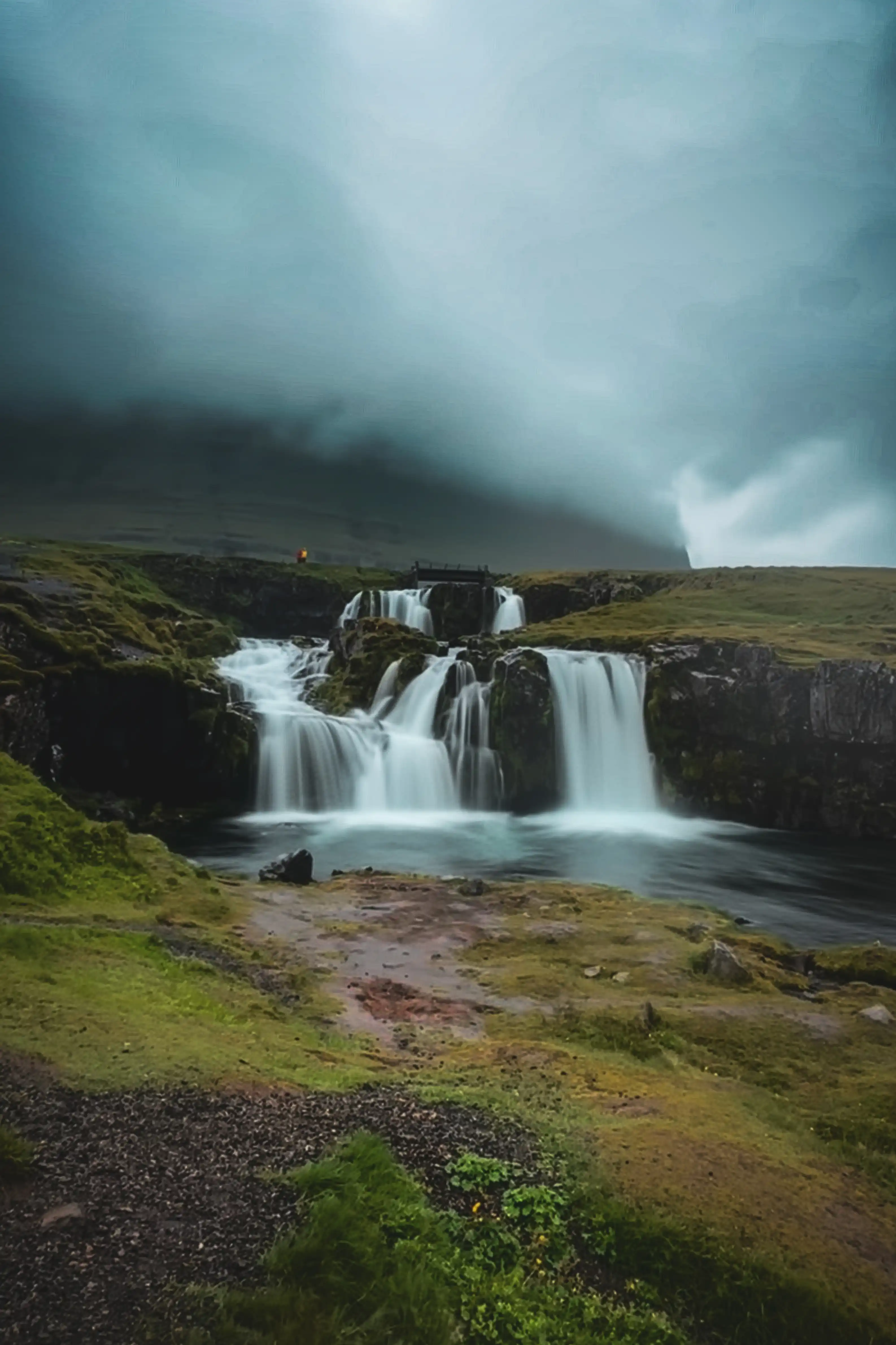 Icelandic waterfall beneath misty low clouds and green hills.