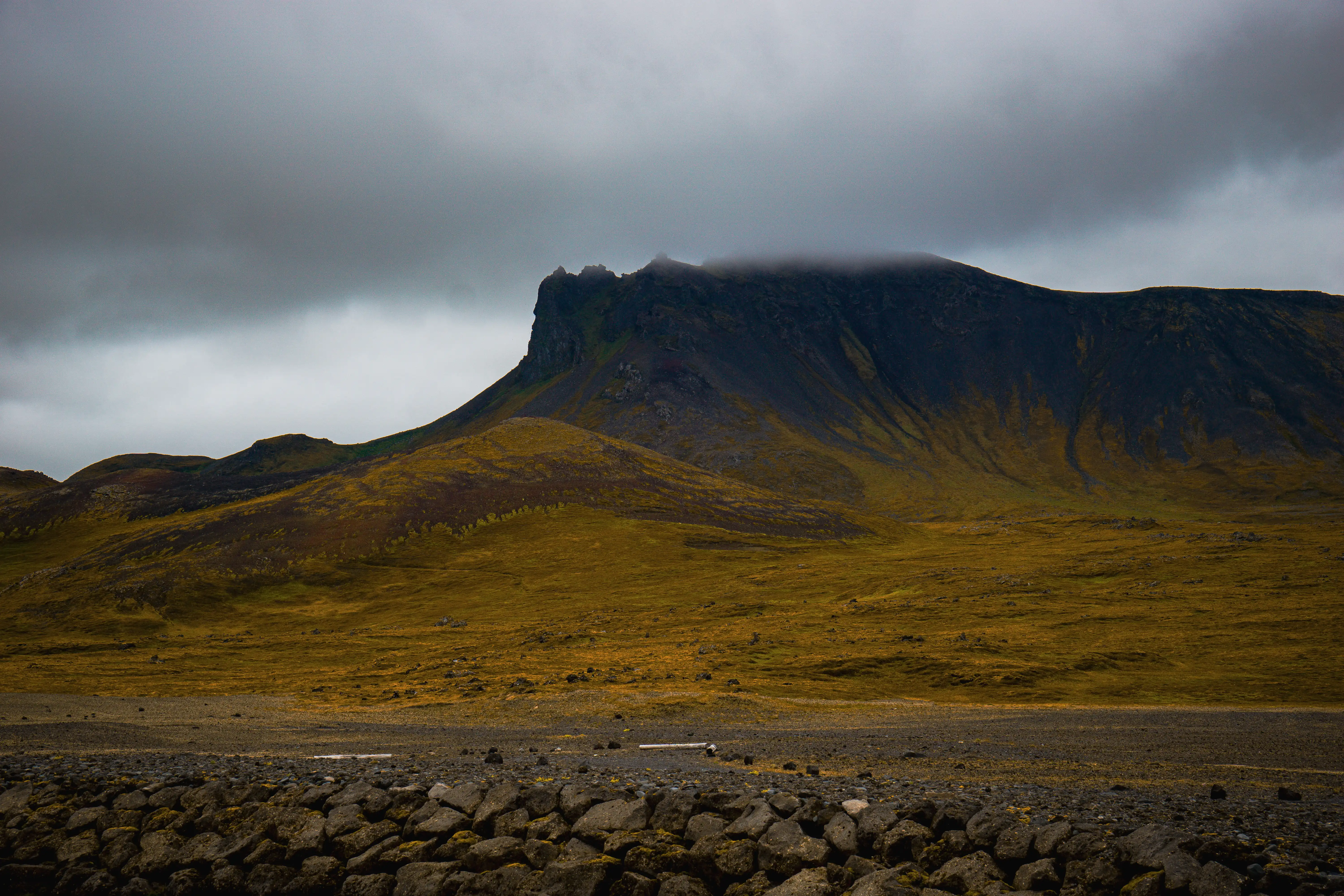 Dark Icelandic mountain under heavy clouds with golden mossy plain below.